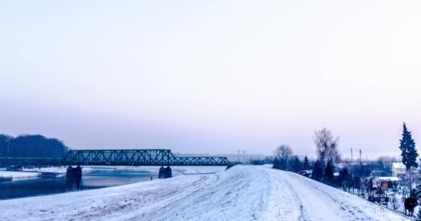 El hombre camina en el río por el puente del ferrocarril de nieve Hiperlapso Titelapse Pequeñas casas entre los árboles Ramas desnudas Árboles Temprano en la noche Invierno Cielo gris — Vídeos de Stock