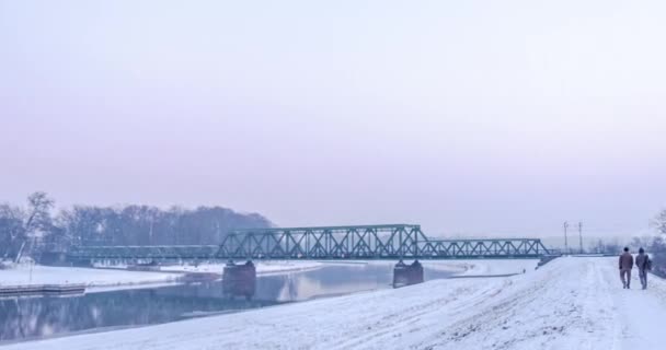 La gente está caminando a lo largo de una orilla del río en tren de nieve se mueve por el puente ferroviario Hiperlapso Titelapse Casas pequeñas Ramas desnudas Árboles Temprano en la noche Invierno — Vídeo de stock