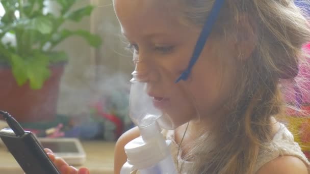 Kid tittar uppmärksamt på telefonens skärm sitter med nebulisator mask på hennes ansikte håller en mobiltelefon unge andas genom inhalator ånga — Stockvideo