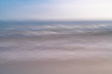 Plaj hareketi soyut kumları bulanıklaştırır ve yumuşak odaklanmış renklerde dalgalar yavaşça kumsala vurur..