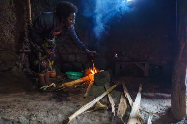 Gasura Ruanda - 8 Eylül 2025; Otantik Kadın dumanlı ateşin üzerine eğilip yeşil kaseden ateşe yemek koyarak geleneksel şekilde pişirmek için küçük karanlık bir kulübede ya da evde.