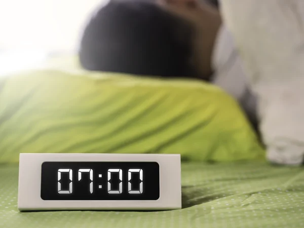 7 en punto en reloj digital con el hombre durmiendo en la cama —  Fotos de Stock