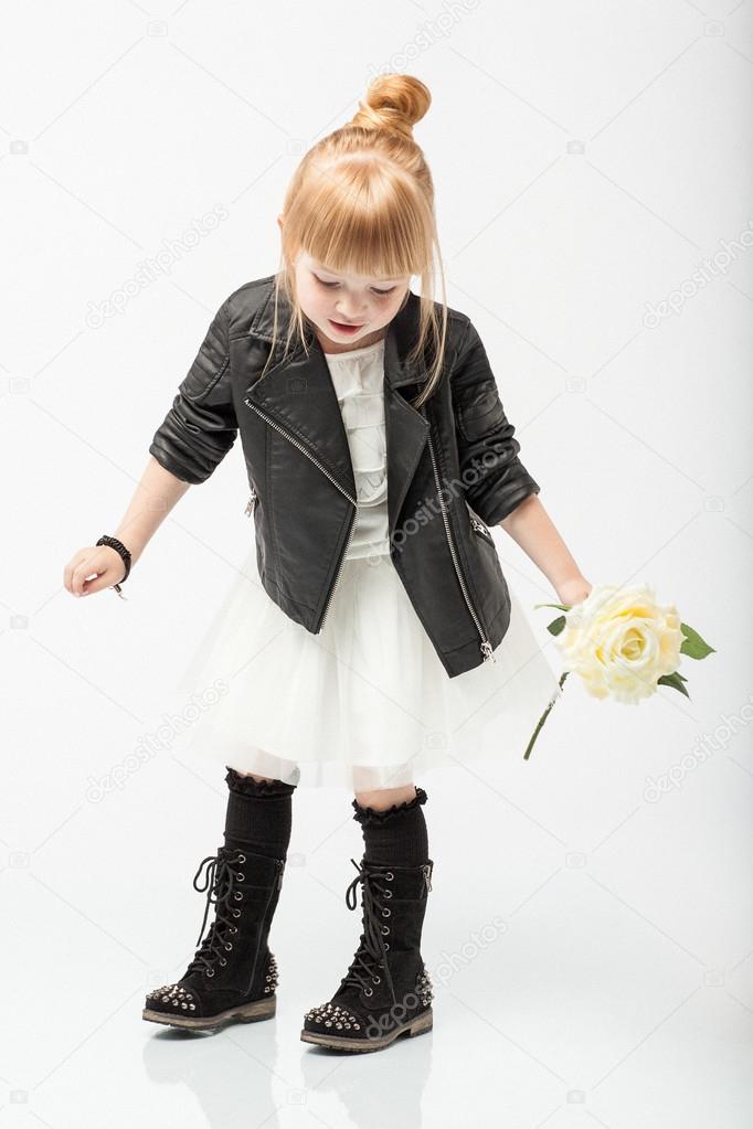 Cute little girl posing in a studio at black biker jacket, white dress and black boots.