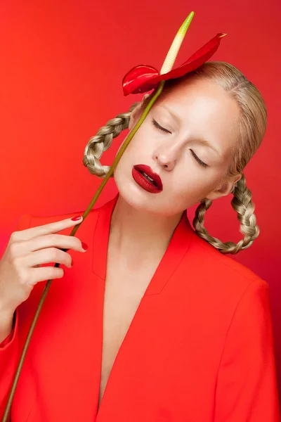 Sensual charming sexy blonde woman in red jacket posing in studio taking red calla on her hand, red background. — Stockfoto