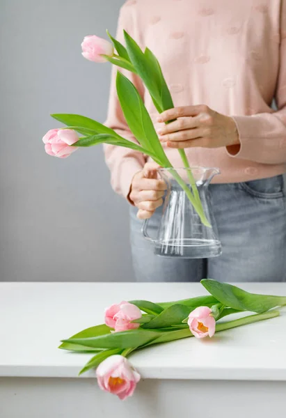 pink tulips with green leaves in a glass vase, a woman holding tulips in her hands