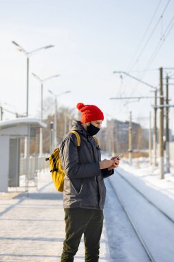 Koyu renk ceketli ve kırmızı şapkalı beyaz bir adam tren istasyonunda tren bekliyor ve akıllı telefonuyla mesaj atıyor.