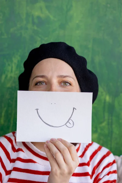 European woman in a black beret holding a sticker with a smiley face, painted smile as a mask on a womans face