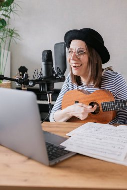 Podcast, müzik içeriği oluşturma. Güzel Avrupalı kadın podcaster, gitar ya da ukulele şapkalı, radyo sunucusu podcast veya içerik kaydediyor, şarkı ve müzik bir kayıt stüdyosunda