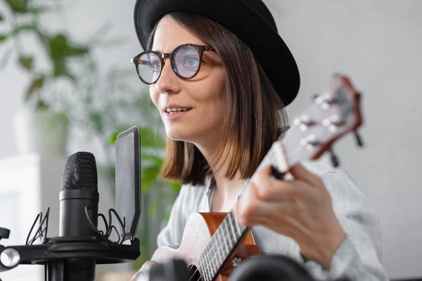 Podcast, creación de contenido de audio musical. hermosa mujer europea podcaster en un sombrero con una guitarra o ukelele, radio anfitrión grabación podcast o contenido, canción y música en un estudio de grabación — Foto de Stock