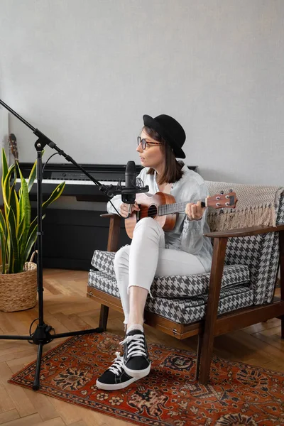 Podcast, music audio content creation. beautiful European woman podcaster in a hat with a guitar or ukulele, radio host recording podcast or content — ストック写真