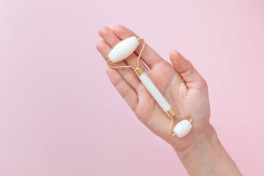 Beyaz kuvars masaj aleti, pembe arka planda bir kadının eli, yüz ve boyundaki deriyi sıkılaştırmak için masaj aleti, kırışıklıkları ve yaşlanma etkilerini yok etmek için.