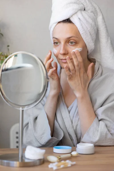 Natural beauty and skin care, beautiful young caucasian woman applies cream on her face, beauty of face and body, skin health and beauty care, woman with a towel on her head in front of the mirror — Stock Fotó