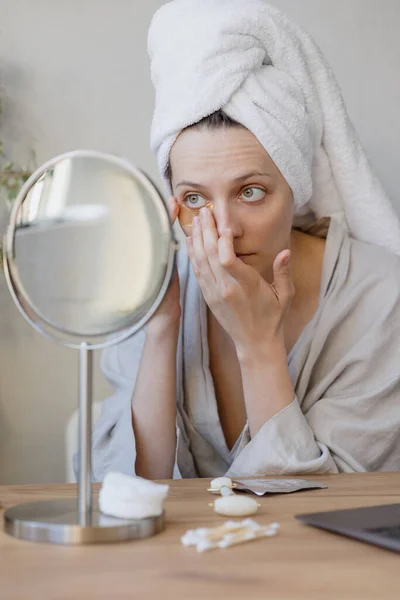 Natural beauty and self-care, Caucasian woman with a towel on her hair in front of the mirror applies moisturizing cream or patches on her face, the woman takes care of her skin, health and beauty — Stock Fotó