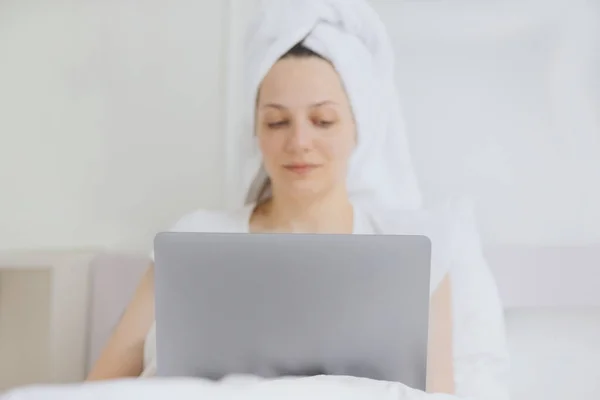 Attraktive europäische Frau in einem weißen Bett mit einem Handtuch auf dem Haar plaudert auf ihrem Handy oder Laptop, entspannen im Urlaub oder auf Reisen — Stockfoto