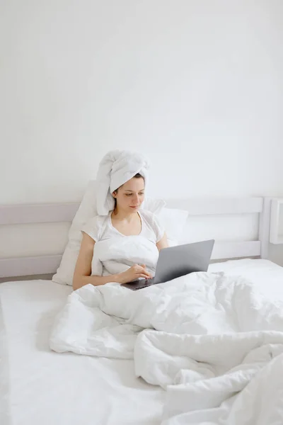 Attraktive europäische Frau in einem weißen Bett mit einem Handtuch auf dem Haar plaudert auf ihrem Handy oder Laptop, entspannen im Urlaub oder auf Reisen — Stockfoto