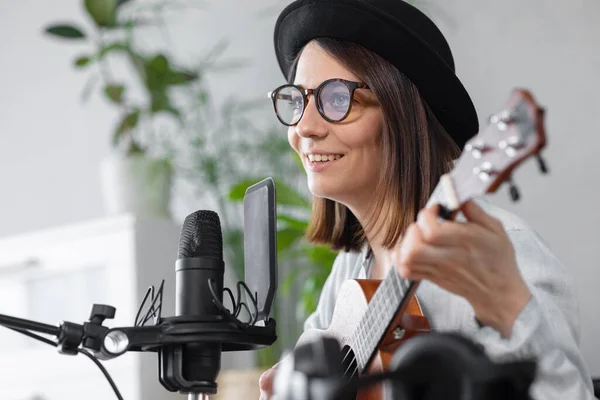 Podcast, creación de contenido de audio musical. hermosa mujer europea podcaster en un sombrero con una guitarra o ukelele, radio anfitrión grabación podcast o contenido, canción y música en un estudio de grabación — Foto de Stock