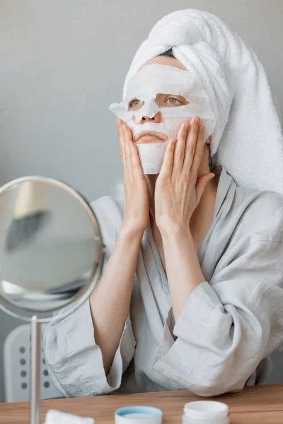 Európai nő ül a tükör előtt, és egy fehér ruha hidratáló maszkot az arcára, bőrápoló és spa kezelések otthon, nő kozmetikus csinál anti-aging arc maszk — Stock Fotó