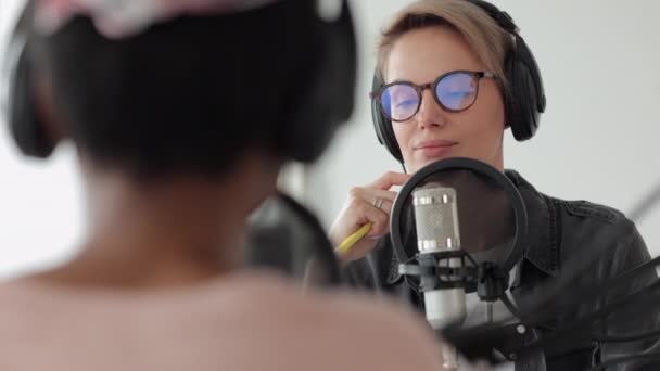 Een Europese vrouw met koptelefoon spreekt in een microfoon, twee vrouwen in een opnamestudio maken audio-inhoud — Stockvideo