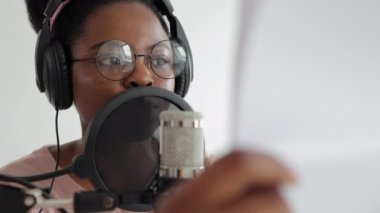 Podcast ve ses içeriği oluşturma, özgüvenli ve güzel bir Afrikalı-Amerikalı kadın mikrofon ve kulaklıkla bir kayıt stüdyosunda veya evinde podcast kaydediyor.