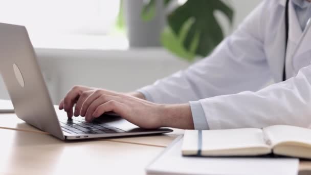 Médico con una bata blanca en su consultorio en una clínica médica, el médico trabaja después de ver a los pacientes en el ordenador escribiendo — Vídeos de Stock