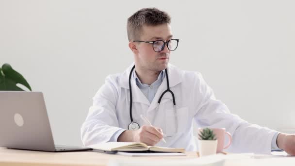 診療所の事務所にある白いコートの医者が日記に記入します医者は患者をコンピューターで見た後に働きます, — ストック動画