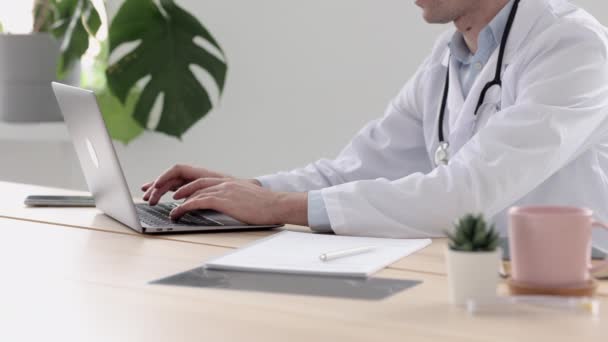 Médico con una bata blanca en su consultorio en una clínica médica, el médico trabaja después de ver a los pacientes en el ordenador escribiendo — Vídeos de Stock