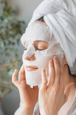 Avrupalı bir kadın yüzüne beyaz bez maskeler, yüz bakımı ve spa tedavileri uyguluyor. Kadın güzellik uzmanı yaşlanma karşıtı yüz maskesi yapıyor.