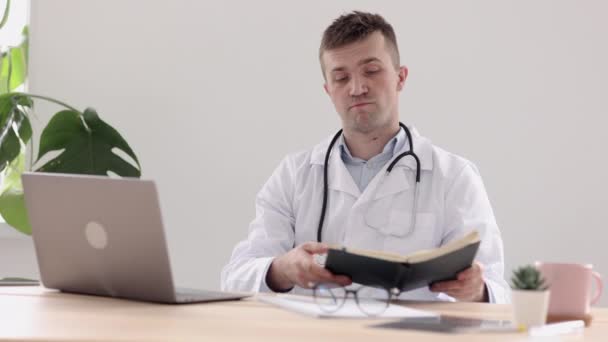 Telemedicina, un médico europeo con una bata blanca y gafas hace una nota en un diario y una anotación en un cuaderno — Vídeos de Stock