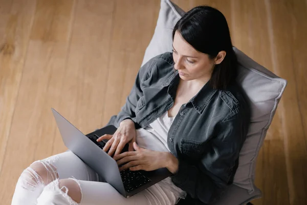 Von oben, europäische Geschäftsfrau in grauem Hemd und weißer Jeans sitzt auf einem Stuhl, arbeitet mit einem Laptop und chattet am Telefon, SMS, surft im Internet oder erledigt Online-Einkäufe — Stockfoto