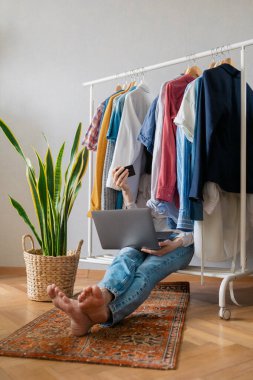 Beyaz Avrupalı kadın gardıropta oturuyor, kıyafetlerin arasında ve internetten alışveriş yapıyor, dizüstü bilgisayarı ve telefonu olan kadın çevrimiçi mağazadan yeni bir elbise ve diğer giysiler sipariş ediyor.