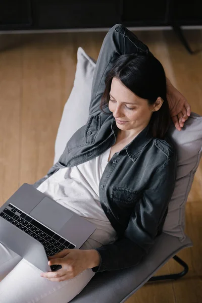 Von oben, europäische Geschäftsfrau in grauem Hemd und weißer Jeans sitzt auf einem Stuhl, arbeitet mit einem Laptop und chattet am Telefon, SMS, surft im Internet oder erledigt Online-Einkäufe — Stockfoto