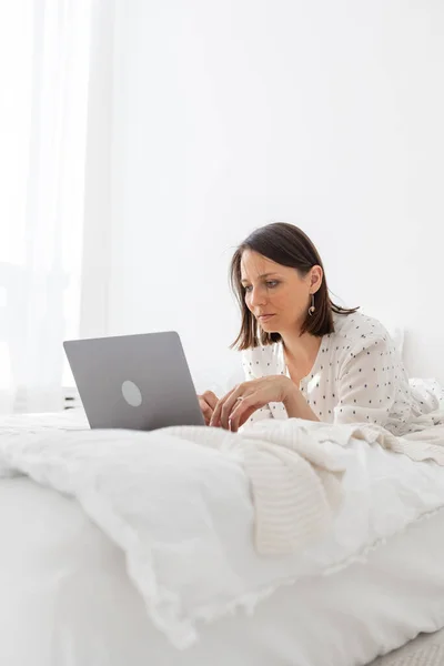 Eine junge und schöne Europäerin liegt auf einem Bett in einem weißen Schlafzimmer mit Laptop und arbeitet — Stockfoto