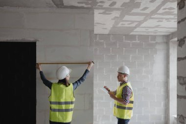 İş planını tartışmak için inşaat alanında beyaz kasklı kadın ve erkek mühendisler ve sarı yelekli adamlar var.