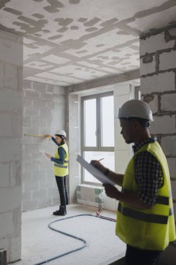 İş planını tartışmak için inşaat alanında beyaz kasklı kadın ve erkek mühendisler ve sarı yelekli adamlar var.