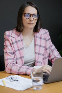Pembe ceketli Avrupalı kendine güveni tam bir iş kadını ve ofisinde laptopuyla çalışıyor.