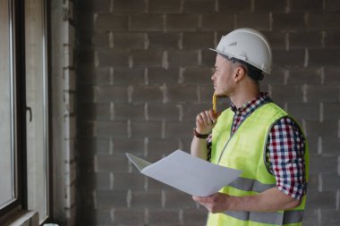 İnşaat alanında beyaz kasklı ve güvenlik yeleği giyen Avrupalı erkek mühendis planı inceliyor.