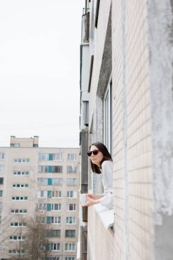 Güneş gözlüklü Avrupalı bir kadın ve bir kupa kahve ya da çayla pencereden dışarı bakıyor ve gülümsüyor.