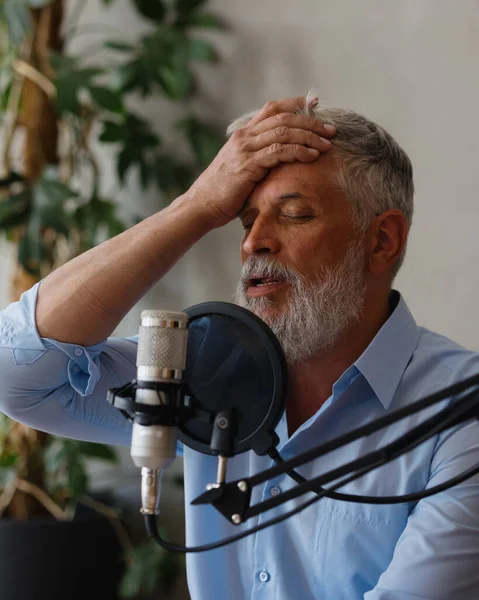 a senior man records audio content or conducts a radio program in a recording studio. a man with gray hair and a beard in the studio with headphones and a microphone