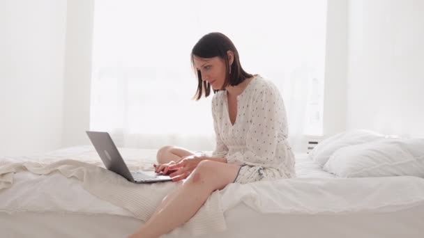 Европейская женщина в белой комнате сидит на кровати и работает на ноутбуке. Кавказская женщина в своем домашнем офисе отправляет сообщение коллегам или партнерам — стоковое видео