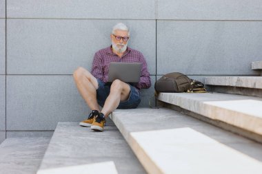 Yetişkin, gri saçlı, sakallı bir adam ofiste oturuyor ve bir dizüstü bilgisayar üzerinde çalışıyor.