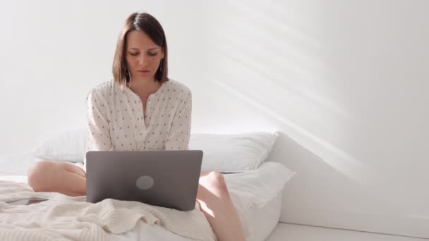 Европейская женщина в белой комнате сидит на кровати и работает на ноутбуке. Кавказская женщина в своем домашнем офисе отправляет сообщение коллегам или партнерам — стоковое видео