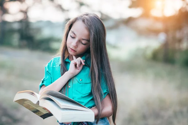 Ładna dziewczynka siedzi na trawie i czytając książkę w — Zdjęcie stockowe