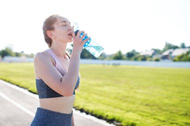 Sıska genç kız stadyumda antrenman yapıyor ve elinde bir şişe su tutuyor.