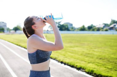 Sıska genç kız stadyumda antrenman yapıyor ve elinde bir şişe su tutuyor.