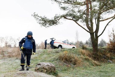 Acil servis çalışanları dışarıda çalışıyor. Üniformalı bir adam metal dedektörüyle çalışıyor.