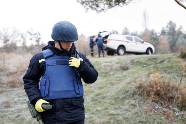 Acil servis çalışanları dışarıda çalışıyor. Üniformalı bir adam metal dedektörüyle çalışıyor.