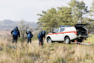 Acil servis çalışanları dışarıda çalışıyor. Üniformalı bir adam metal dedektörüyle çalışıyor.