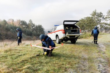 Acil servis çalışanları dışarıda çalışıyor. Üniformalı bir adam metal dedektörüyle çalışıyor.