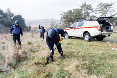 Acil servis çalışanları dışarıda çalışıyor. Üniformalı bir adam metal dedektörüyle çalışıyor.
