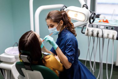 Genç bir kadın dişçi koltuğuna oturur ve dişçi hastanın dişlerini dikkatlice muayene eder. Dişçi, klinikteki bir hastaya danışır.